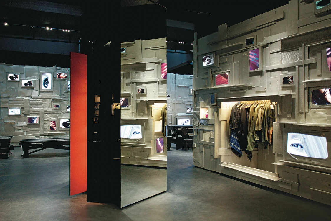 A modern store with structured walls that incorporate screens, niches, and clothing racks. Mirrors and red accents create design highlights in the elegantly illuminated room.