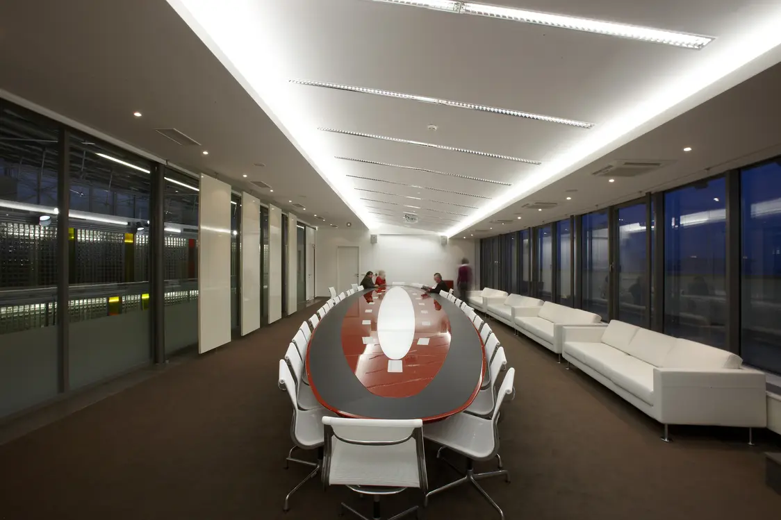 Une salle de conférence moderne avec une longue table ovale, des chaises blanches et une rangée de canapés blancs le long de la fenêtre. Le plafond est équipé de luminaires LED linéaires qui assurent un éclairage clair et homogène.