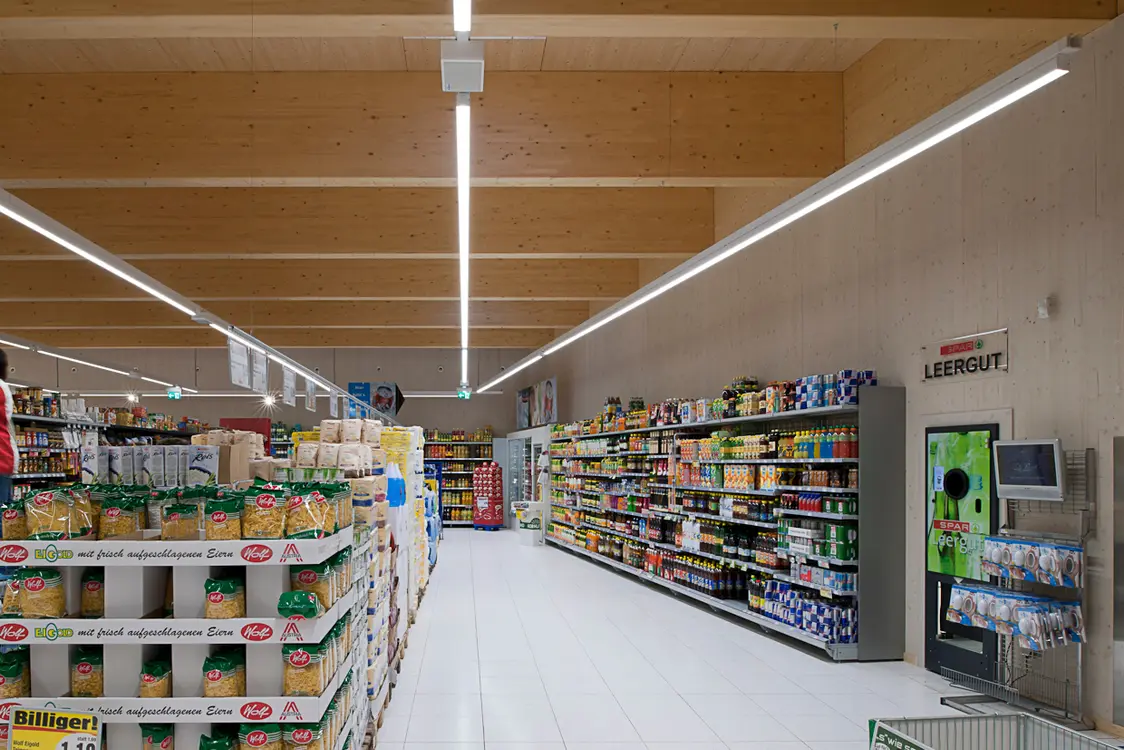 Un moderno supermercato con un soffitto in legno e lunghi apparecchi LED che garantiscono un'illuminazione intensa e omogenea. Gli scaffali sono disposti in modo chiaro e offrono un vasto assortimento di prodotti.