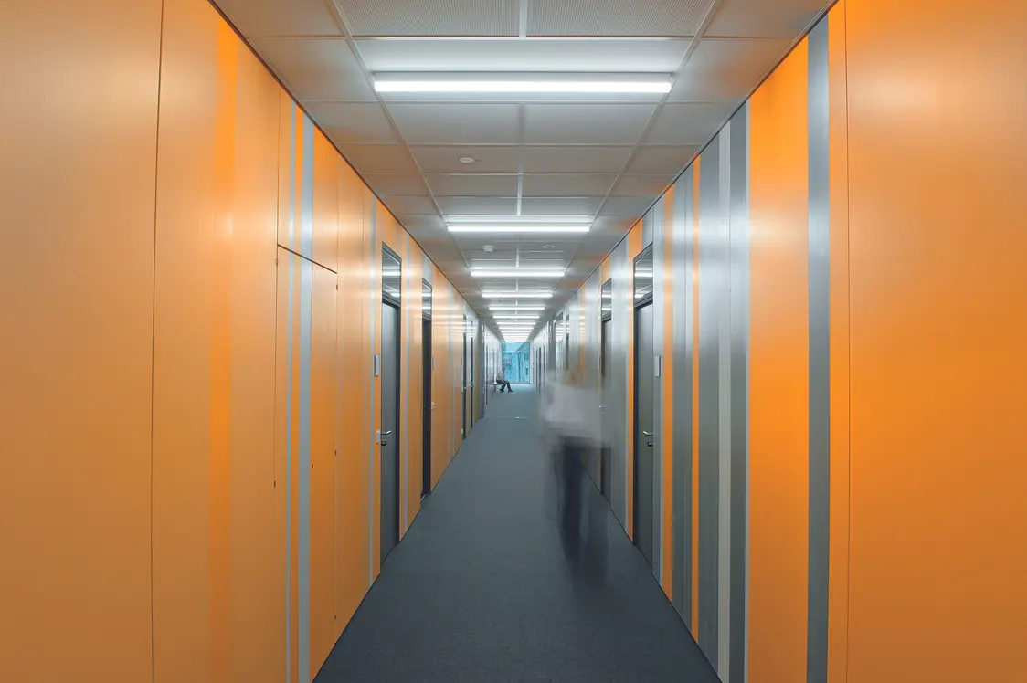 Un long couloir avec des murs à rayures orange et grises, uniformément éclairé par des luminaires au plafond. Une personne se déplace indistinctement dans le couloir