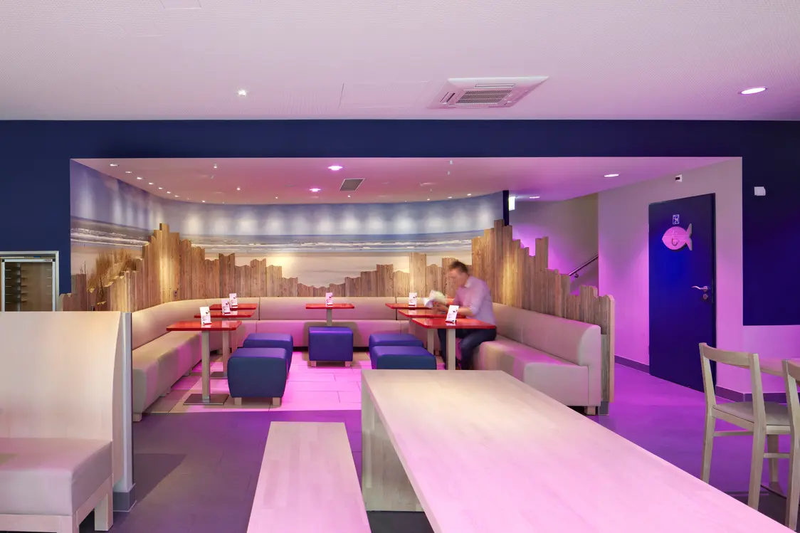 Une cantine moderne avec des meubles en bois clair, des banquettes rembourrées et un éclairage d’accentuation violet. La décoration murale montre un horizon stylisé, et de grandes tables proposent de la place pour des groupes.