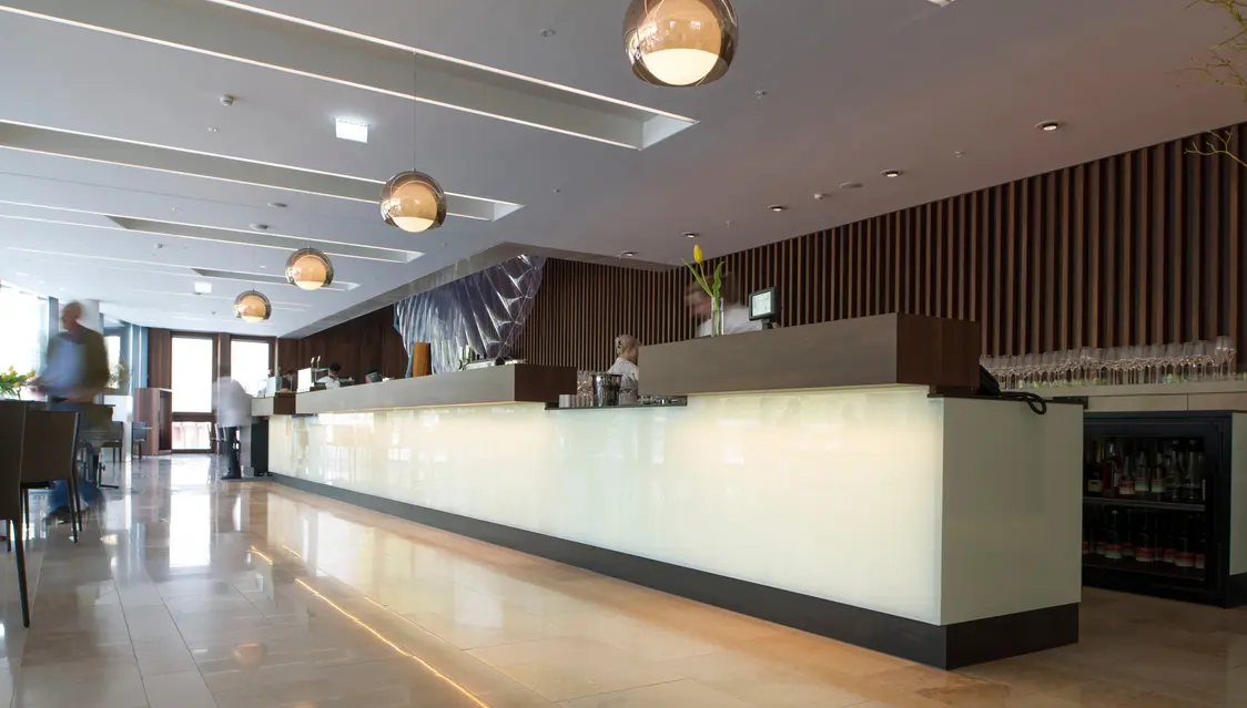 Eine Hotelrezeption mit einer langen, beleuchteten Theke aus Milchglas. Die Decke ist mit eleganten Pendelleuchten ausgestattet, und der Raum wirkt durch die reflektierenden Fliesen hell und einladend.
