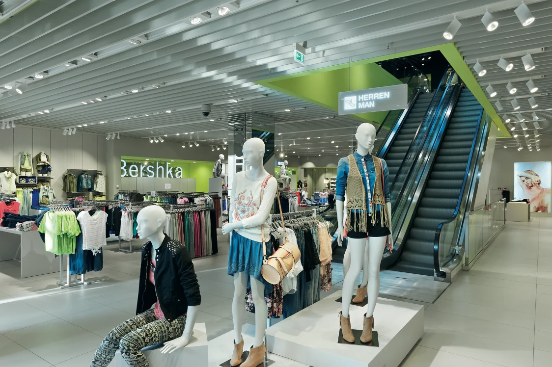 Ampio spazio del negozio con manichini e scala mobile; la soluzione di illuminazione con numerosi faretti sul soffitto mette in risalto i capi di moda e la struttura del locale.