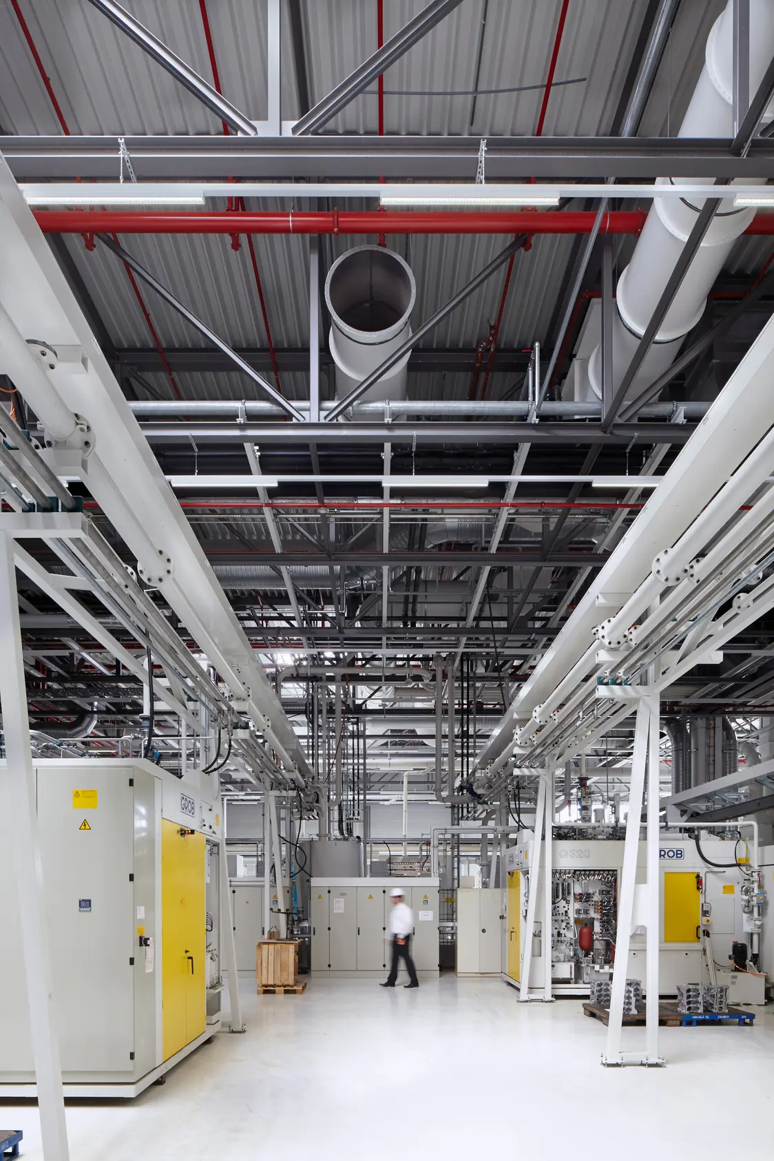 Un hall de production moderne et lumineux avec câblage visible au plafond et de grandes machines. À l’arrière-plan, une personne traverse un espace vaste et bien organisé.