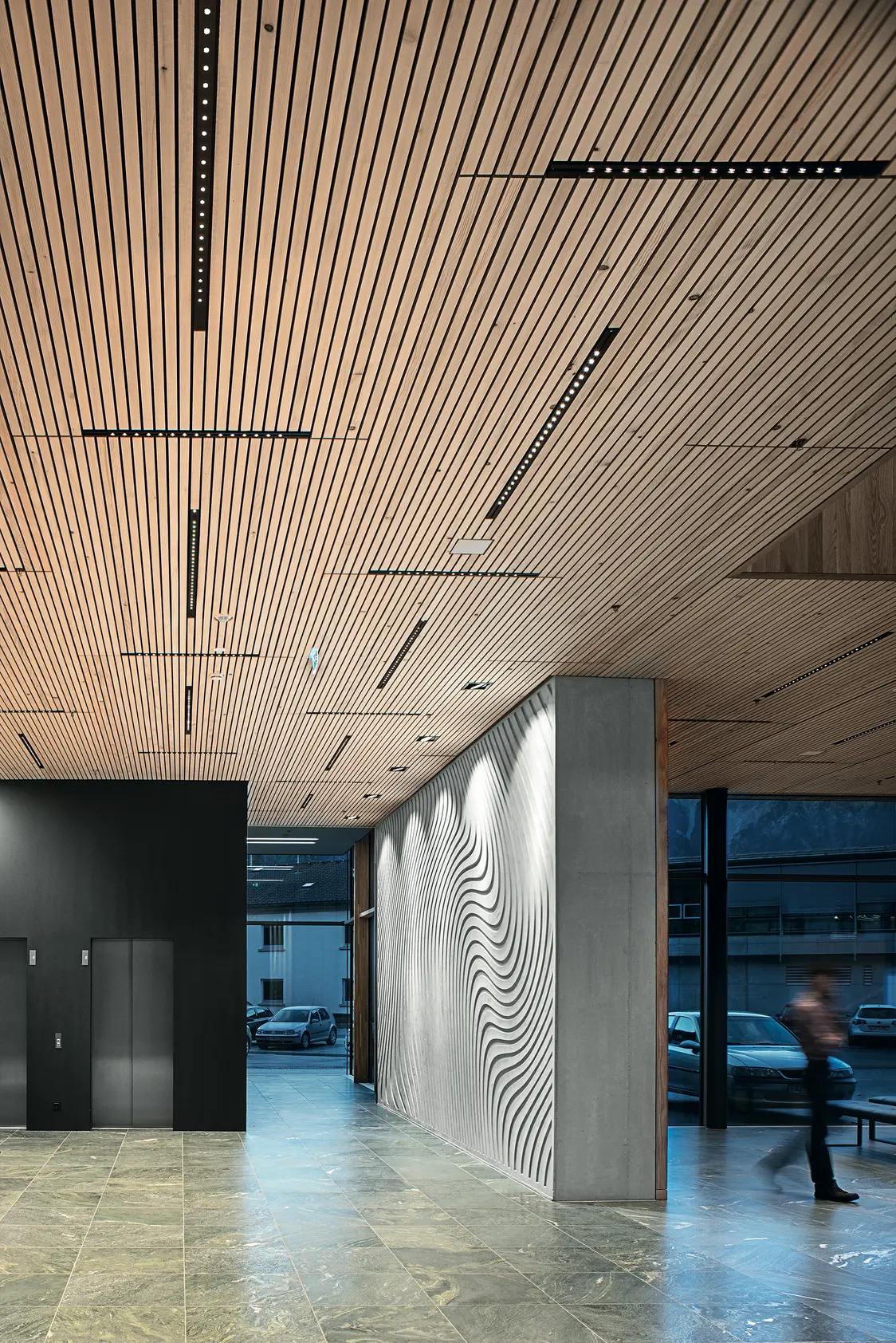 Moderner Eingangsbereich mit heller Holzlamellendecke, eingelassenen LED-Leuchten und einer Wand mit wellenförmigem Muster. Der Raum wirkt offen und großzügig, mit großen Fenstern und Natursteinboden.