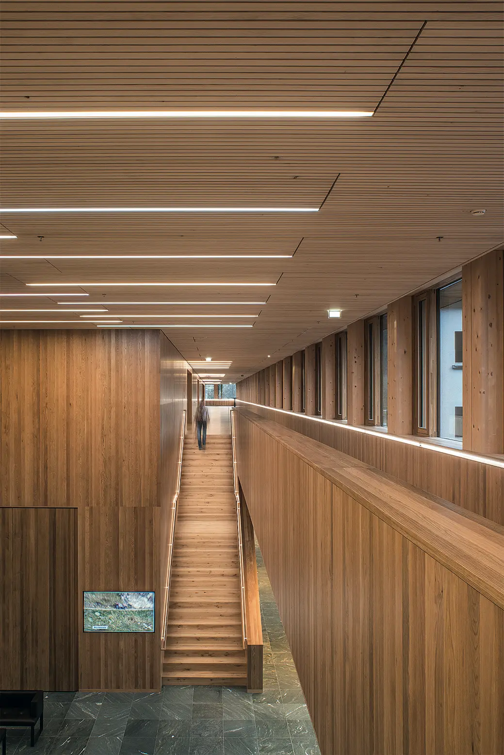 Flur mit Holzverkleidung und Holzlamellendecke, integrierten LED-Lichtbändern sowie einer zentralen Treppe. Große Fenster auf der rechten Seite sorgen für viel Tageslicht und eine helle, freundliche Atmosphäre.