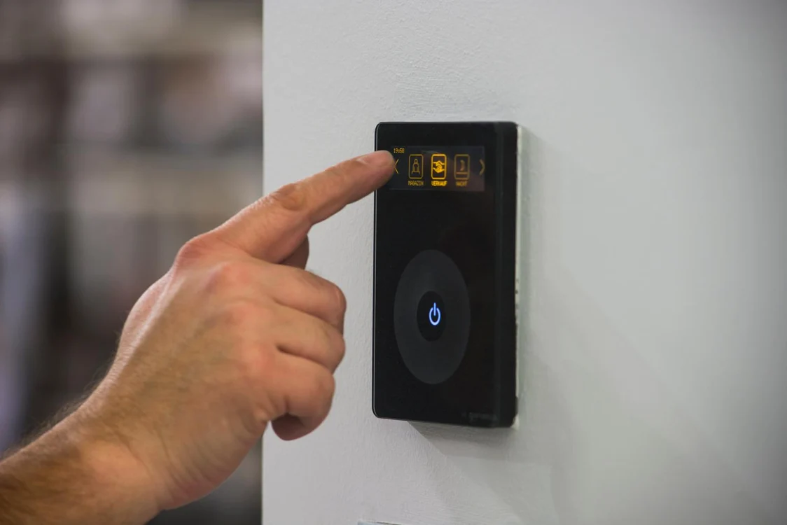 Une main sélectionne des modes d’éclairage sur le panneau de commande LITENET fixé à un mur blanc ; l’arrière-plan est flou et chaleureux.