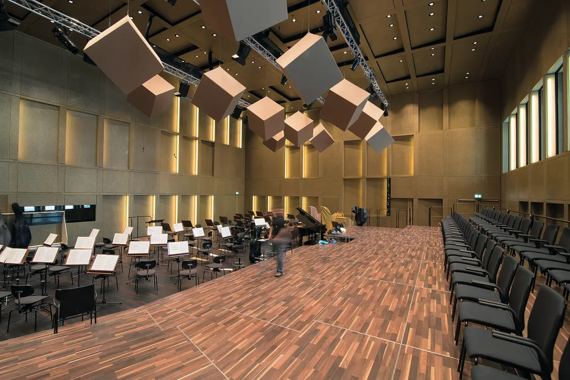Ein Konzertsaal mit Holzfußboden und modernen Akustikelementen, die von der Decke hängen. Die Bühne ist hell beleuchtet, und der Raum bietet Sitzplätze auf beiden Seiten.