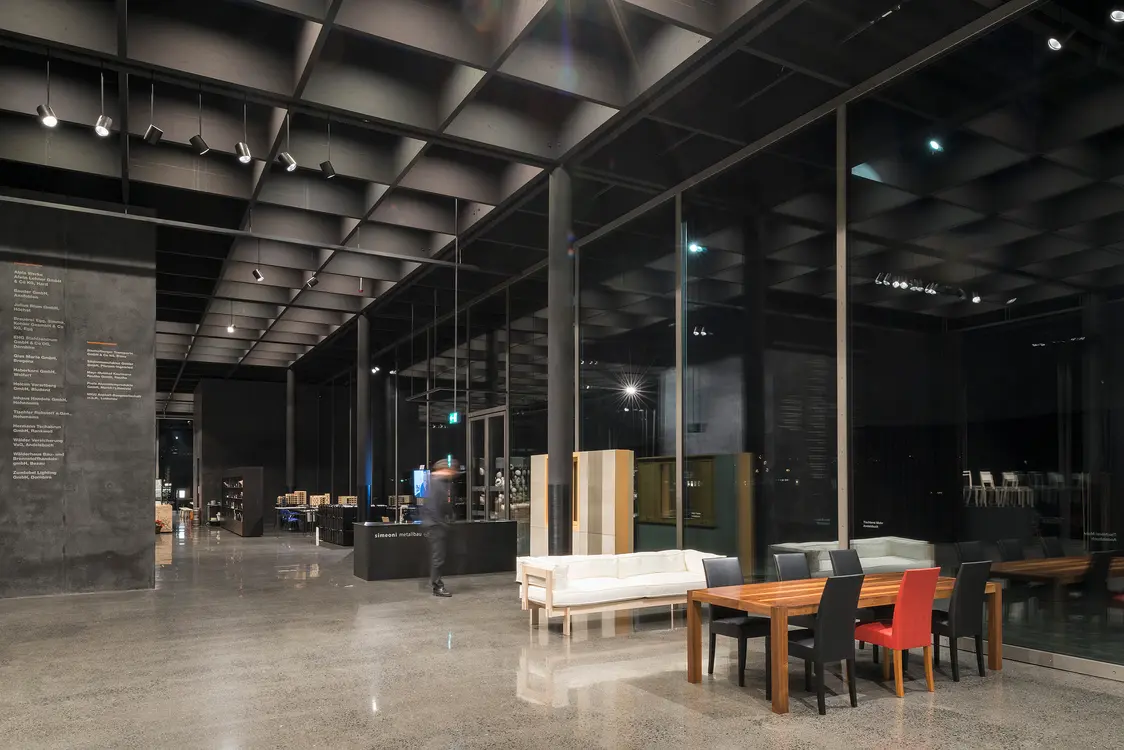 Spazio espositivo moderno con soffitto a doghe scuro e grandi vetrate che creano un ambiente aperto e spazioso. La combinazione di pavimento levigato, mobili eleganti e illuminazione mirata crea un'atmosfera elegante e accogliente.