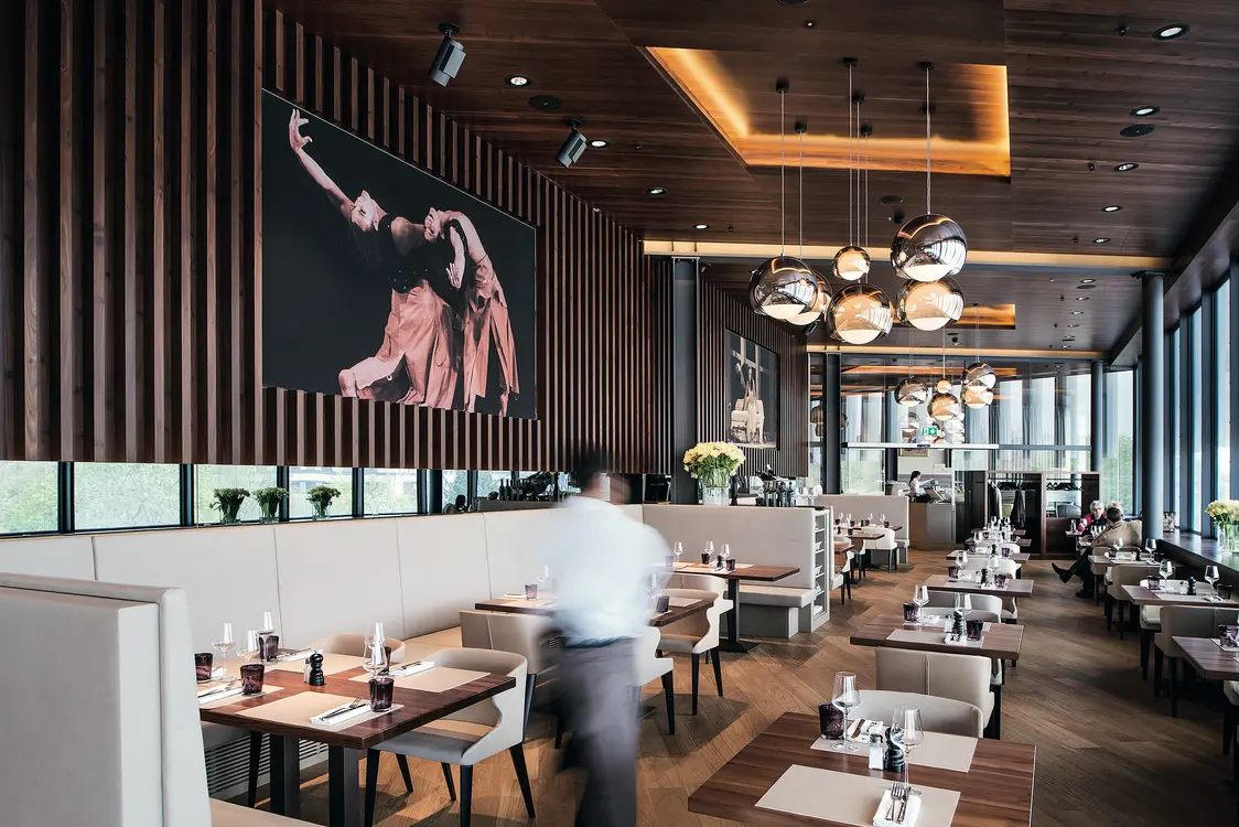 Ein stilvolles Restaurant mit Holzvertäfelung an den Wänden und einer warmen Beleuchtung durch moderne Pendelleuchten. Die Tische sind elegant gedeckt, und ein großes Kunstwerk mit einem Tänzer ziert die Wand.