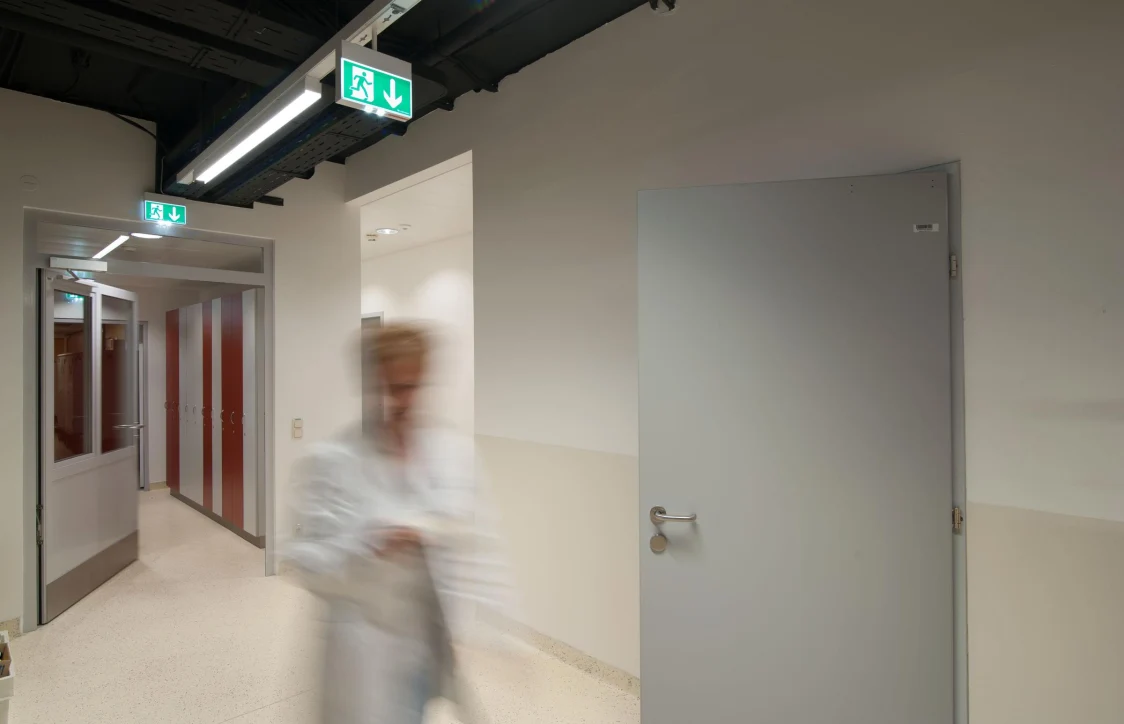 The CROSSIGN luminaire offers guidance in a hospital corridor with open doors, smooth walls, and a blurred person in the foreground.