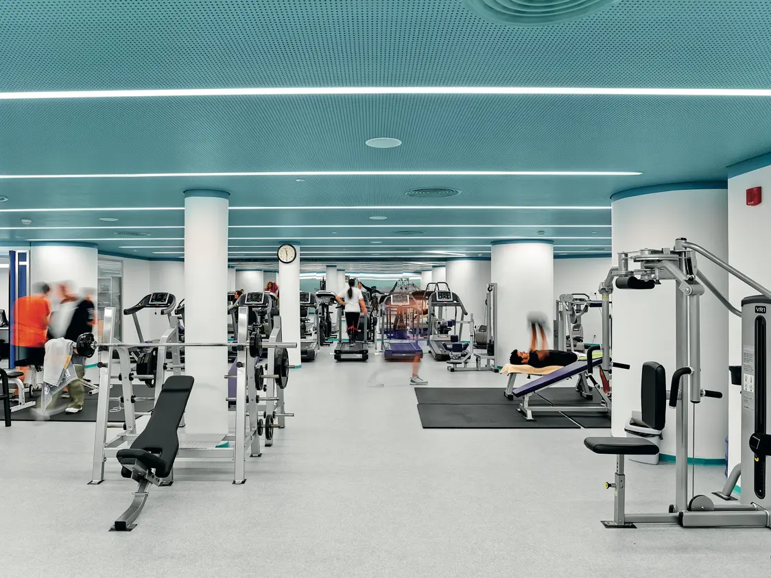 A bright gym with a turquoise ceiling, white columns, and numerous fitness machines. Several individuals are exercising on the equipment or moving through the space.