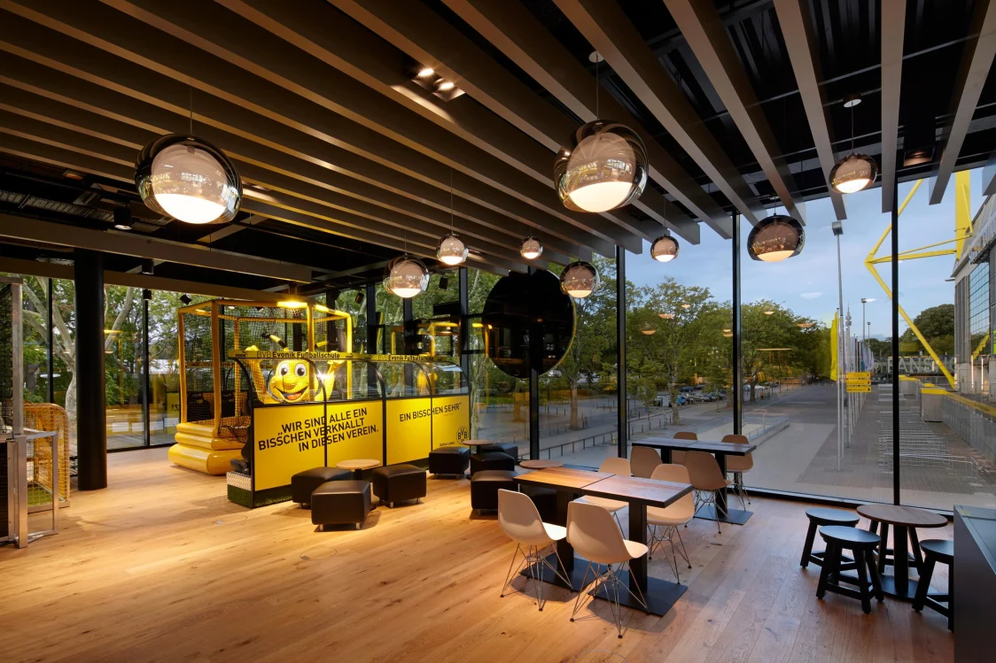 Ein modern gestalteter Innenraum mit großen Glasfronten, hängenden Kugelleuchten und einer gelben Ausstellungsinstallation. Der Raum kombiniert industrielles Design mit gemütlicher Atmosphäre und bietet Sitzgelegenheiten aus Holz und Metall.