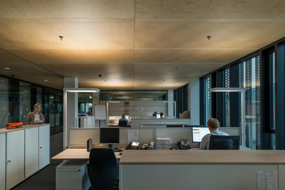 Ein modernes Großraumbüro mit mehreren Arbeitsplätzen, an denen einige Personen am Computer arbeiten. Große Fenster lassen viel Tageslicht in den Raum.