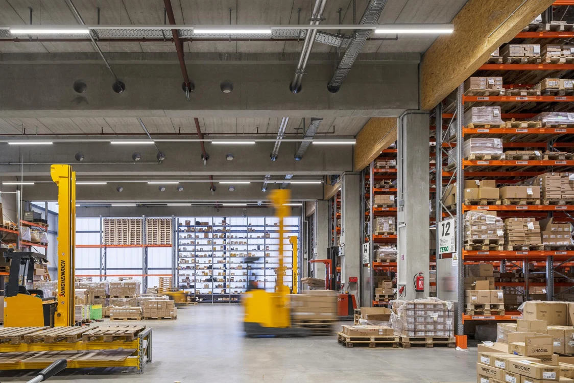 Ein großes Lager mit hohen Regalen voller Kartons und Paletten, in dem ein gelber Gabelstapler Waren transportiert. Die Halle ist hell beleuchtet und wirkt ordentlich organisiert.