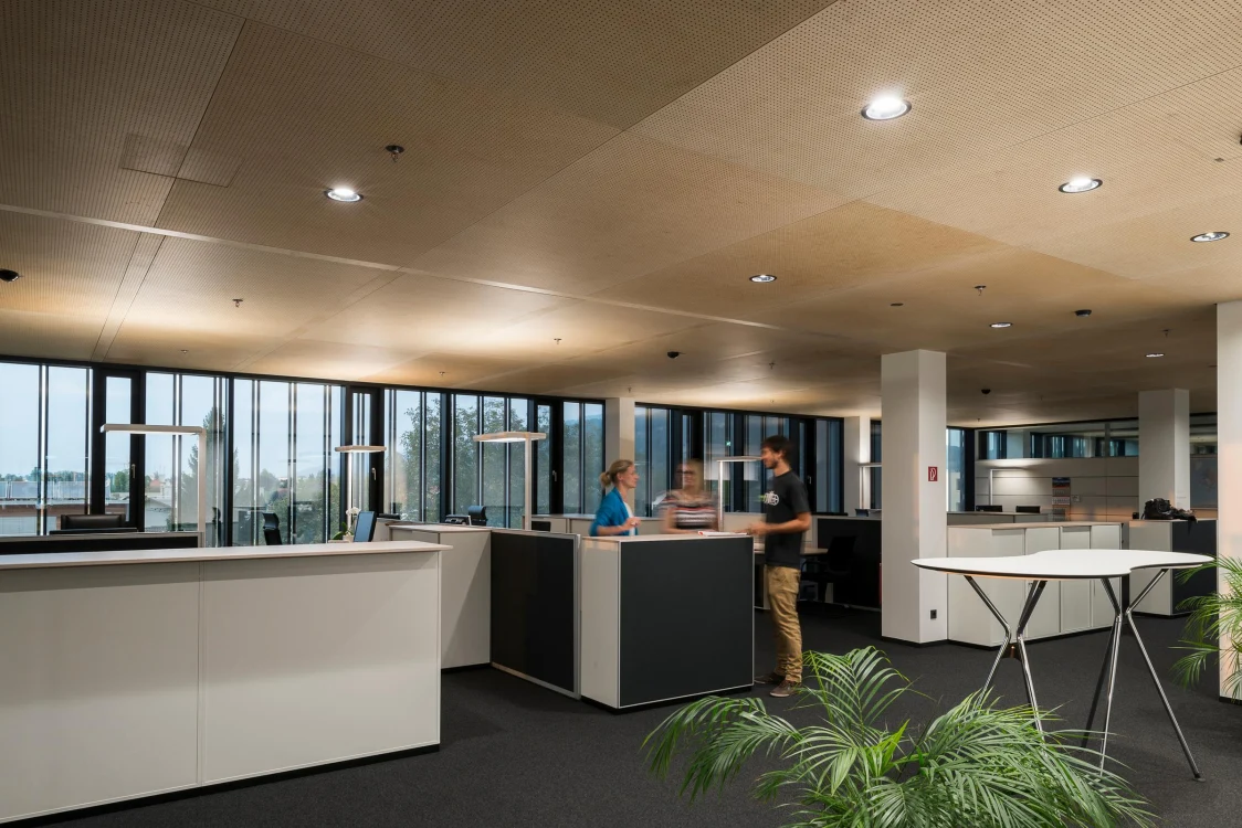 Ein modernes, offen gestaltetes Büro mit großen Fenstern und mehreren Arbeitsplätzen. Zwei Personen unterhalten sich an einem Empfangstresen, während im Hintergrund weitere Schreibtische zu sehen sind.