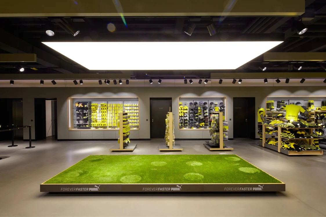 Ein modern gestaltetes Geschäft mit einer großen, leuchtenden Deckenfläche und einer zentralen grünen Kunstrasenplattform. Im Hintergrund sind Regale mit gelb-schwarzen Fanartikeln und Sportzubehör zu sehen.