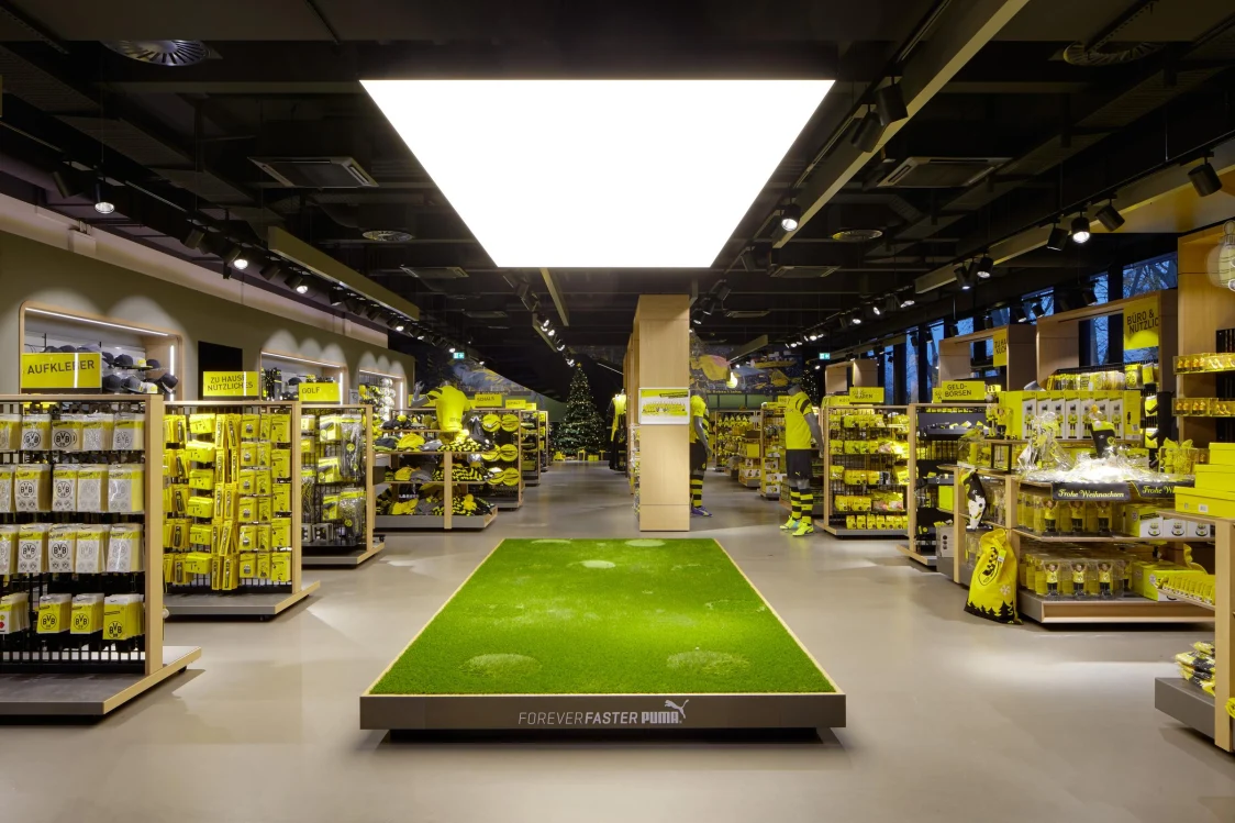 Ein Fanartikelgeschäft mit schwarz-gelber Einrichtung, beleuchtet durch eine große quadratische Deckenleuchte. Im Zentrum befindet sich eine grüne Kunstrasenfläche, umgeben von Regalen mit Merchandise-Produkten.