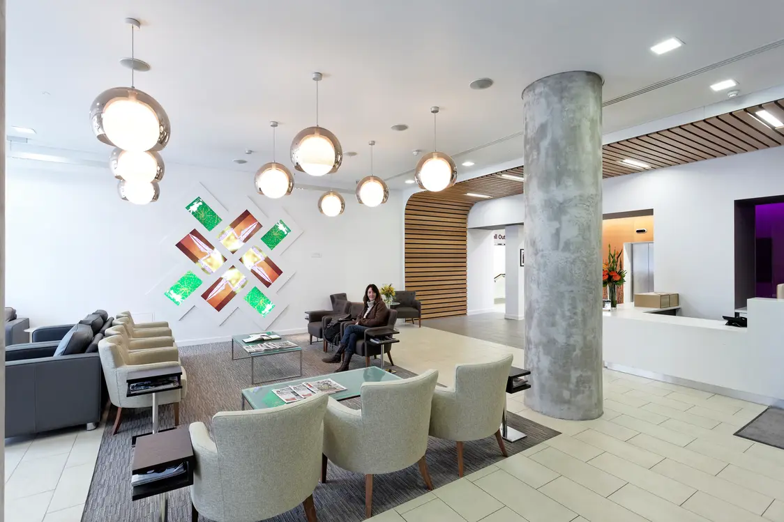 A bright, modern waiting area with comfortable armchairs and round pendant luminaires. A colorful, geometric art installation is hanging on the wall.