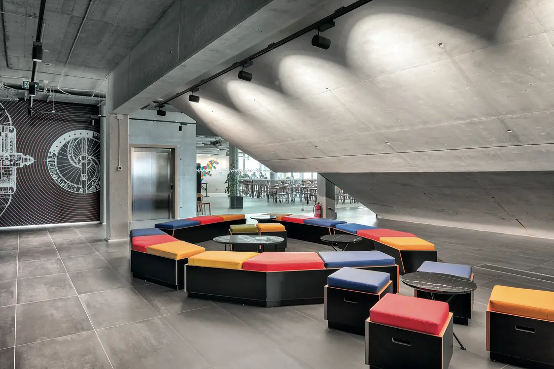 Moderner Innenraum mit farbenfroher Sitzlandschaft unter einer schrägen Betondecke, akzentuiert durch gerichtete Deckenstrahler. Die Lichtlösung betont Materialien und schafft eine zonierte, einladende Atmosphäre im Aufenthaltsbereich.