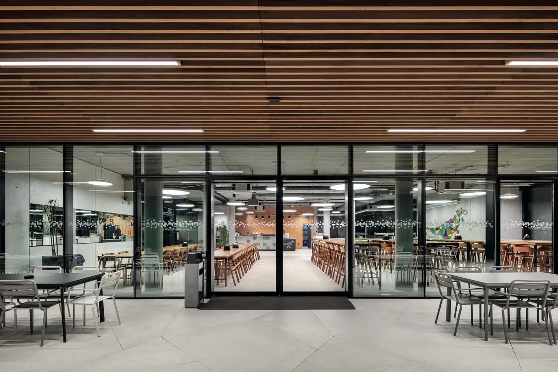 Einladender Blick in die beleuchtete Kantine mit Glasfassade, Holzdecken und klarer Möblierung. Das lineare Lichtkonzept strukturiert die Räume und schafft visuelle Verbindung zwischen Innen- und Außenbereich.