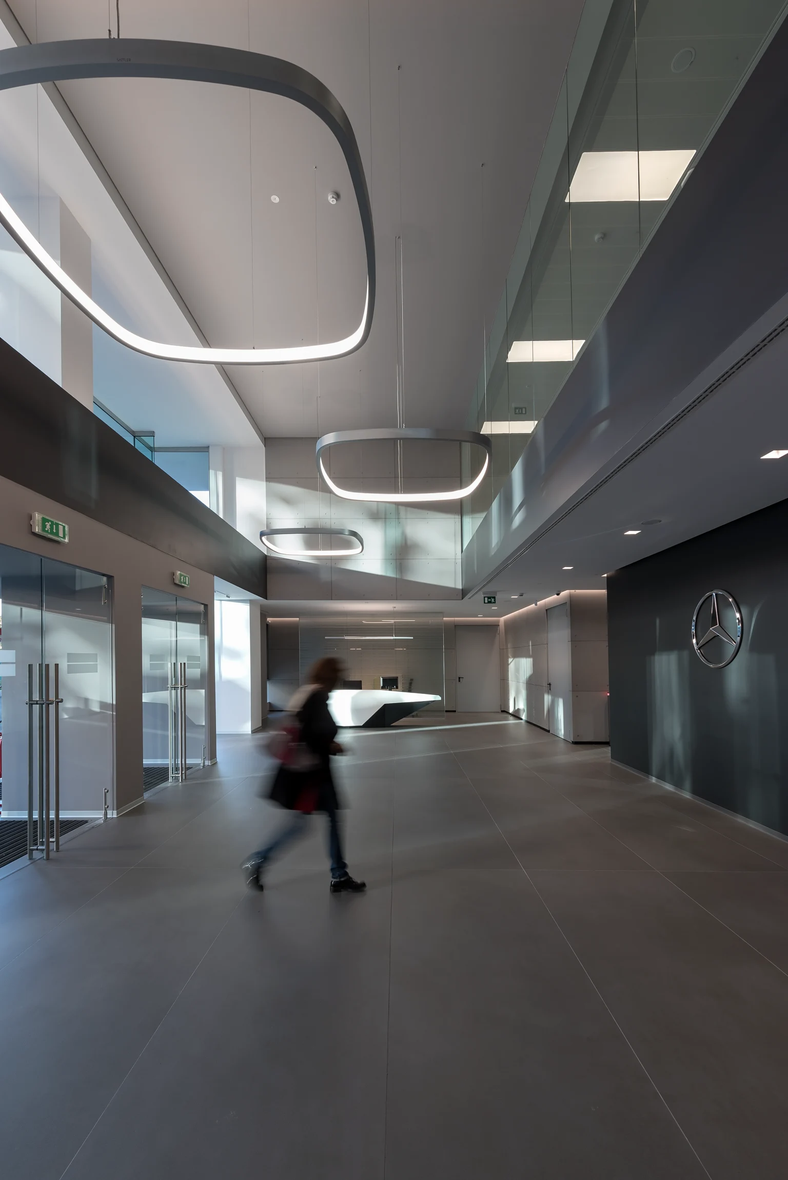 Un hall de bureau moderne avec de hauts plafonds, de grands luminaires géométriques et un logo Mercedes-Benz sur le mur. Une personne traverse la spacieuse entrée au décor minimaliste.
