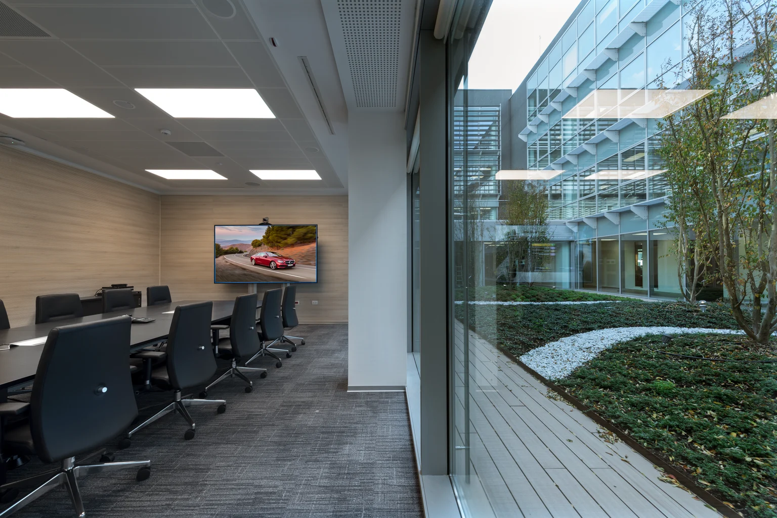 Salle de conférence lumineuse avec une longue table et des chaises de bureau, dotée d’une grande baie vitrée donnant sur un patio avec un aménagement paysager. Un écran sur le mur montre une Mercedes-Benz.