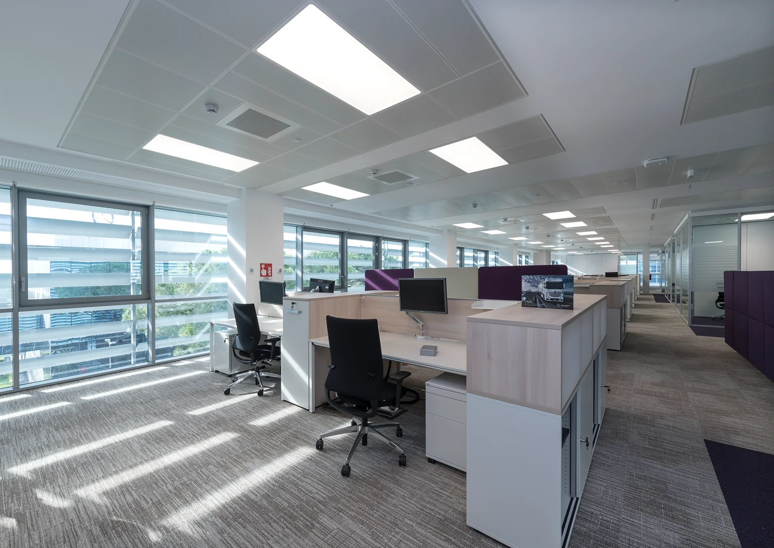 Bureau moderne à l’aménagement ouvert avec de longues rangées de bureaux, des cloisons violettes et une salle de réunion vitrée avec un mobilier minimaliste. Ambiance lumineuse et claire avec des panneaux LED en guise d’éclairage.