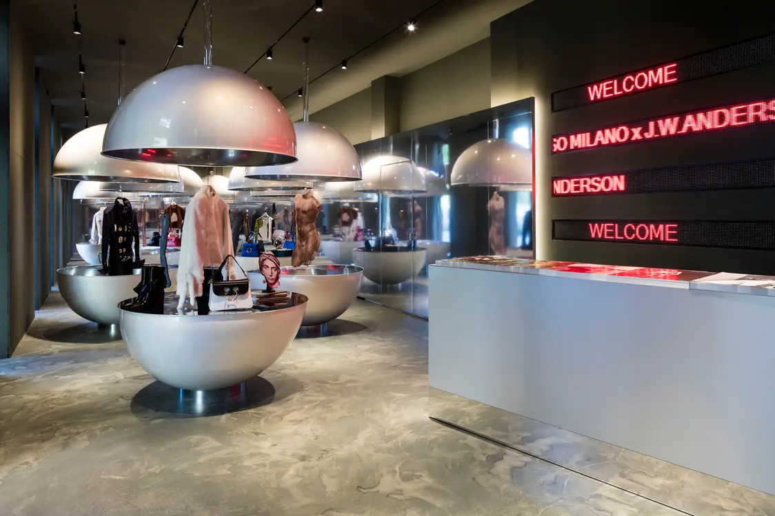 Modern fashion showroom with round display tables under large dome lights, featuring clothing, accessories, and mannequins. A sleek reception desk stands beside a wall with red LED “WELCOME” signage.