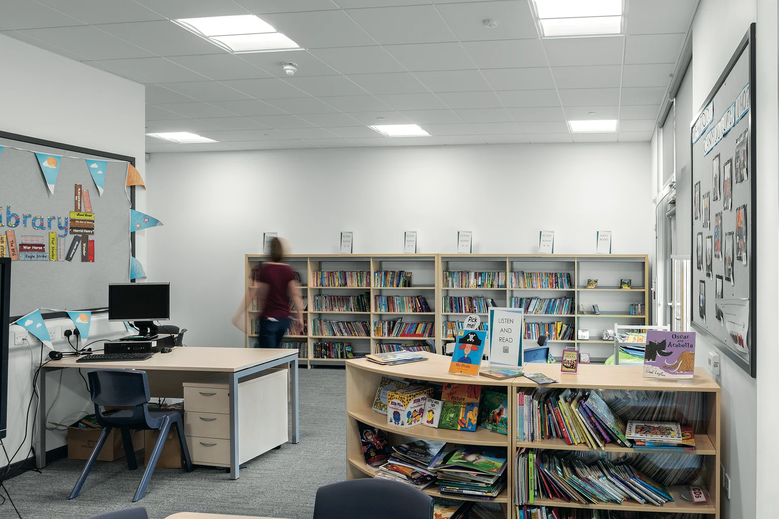 Eine moderne Schulbibliothek mit Bücherregalen, einem gebogenen Display und einem Schreibtisch mit Computer. Der Raum ist gut beleuchtet mit Deckenleuchten und bietet ein sauberes, organisiertes Layout, das Schüler erkunden und zum Lernen nutzen können.