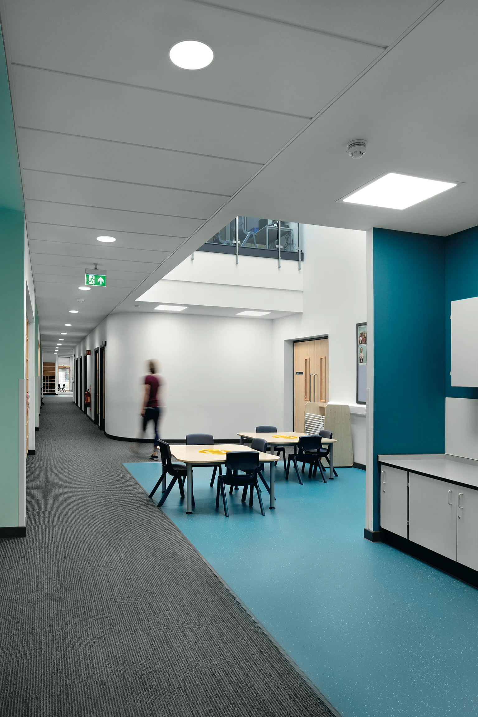 Ein heller und moderner Schulkorridor mit einer Mischung aus Teppich- und Vinylböden, weißen Wänden und türkisfarbenen Akzenten. Der Raum umfasst runde Tische mit Stühlen für gemeinsame Aktivitäten. Er wird von Deckeneinbauleuchten beleuchtet und hat eine sichtbare Treppe, die zur oberen Ebene führt.