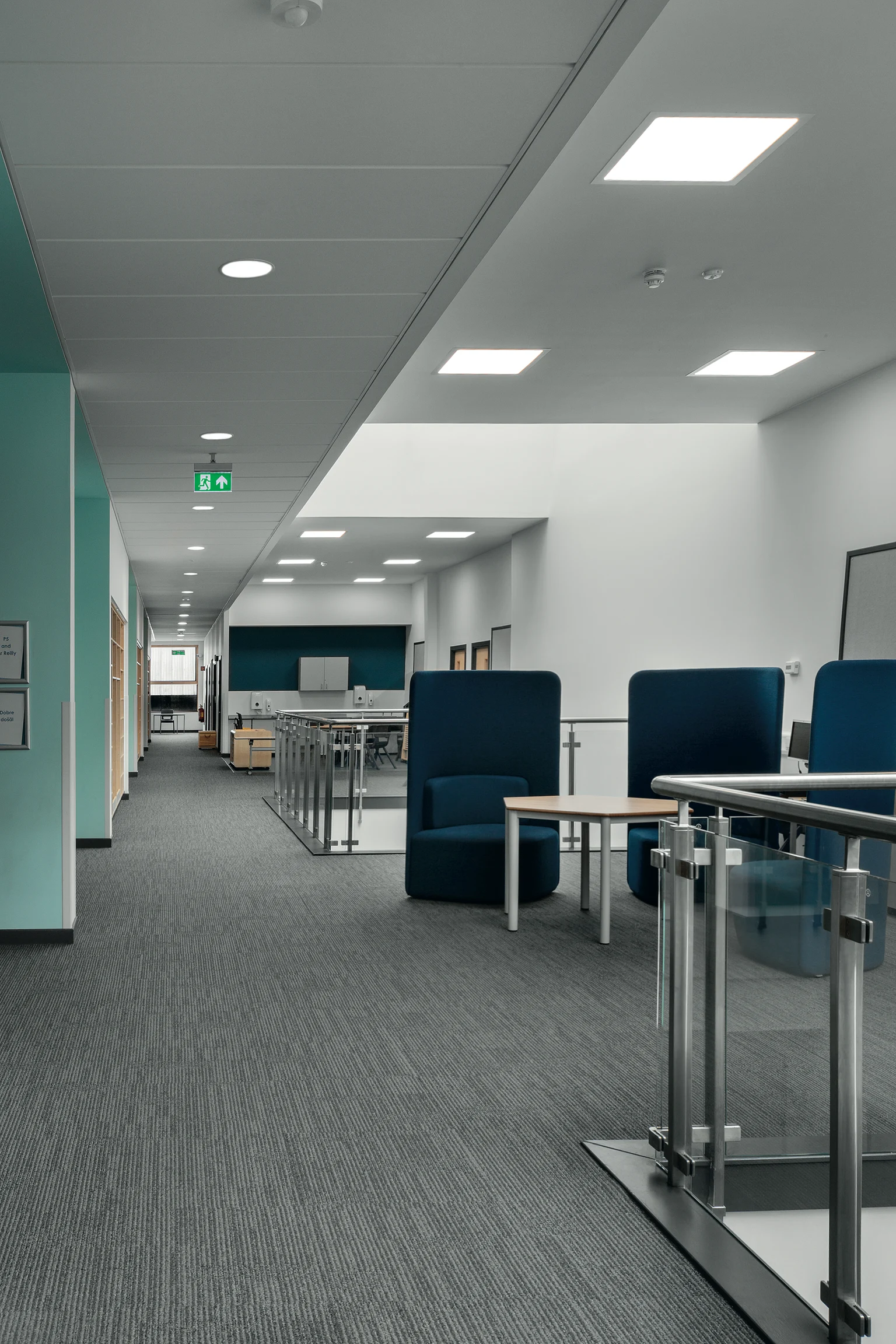 Ein moderner Schulkorridor mit Teppichboden, weißen Wänden und türkisfarbenen Akzenten. Der Raum umfasst Sitznischen mit blauer Polsterung, Schreibtische für gemeinschaftliches Arbeiten und Deckeneinbauleuchten für eine helle Ausleuchtung.