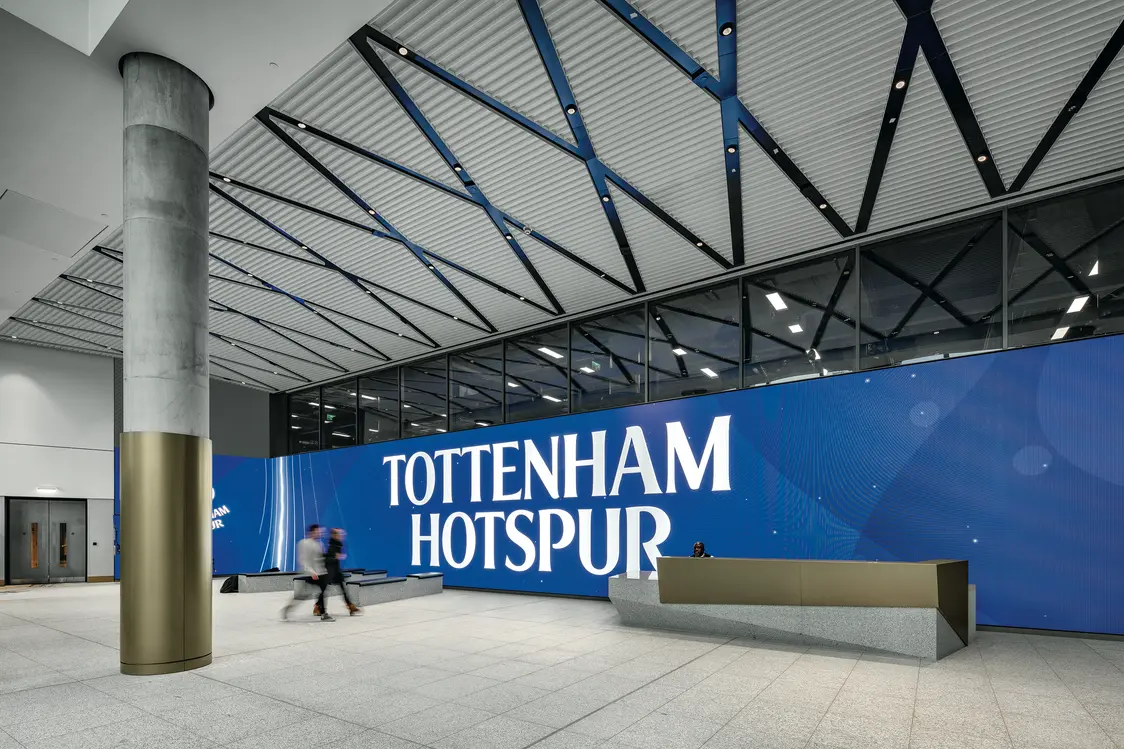 Ein großzügiger Eingangsbereich mit einer großen blauen Wand, auf der „Tottenham Hotspur“ steht, und einem modernen Empfangstresen; die Lichtlösung mit linearen Deckenleuchten betont die Weite und Modernität des Raumes.