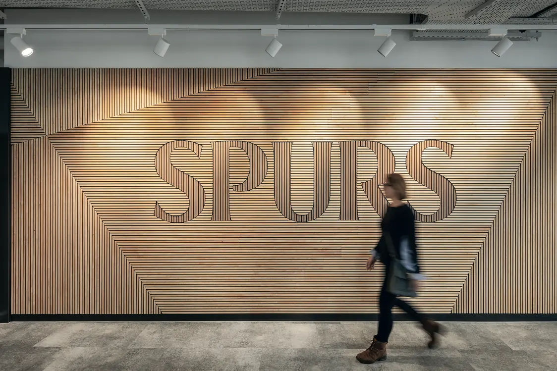 Wand mit Holzlamellen und großem „SPURS“-Schriftzug, akzentuiert durch gerichtete Spotlights an der Decke, die für eine warme und strukturierte Lichtlösung sorgen.