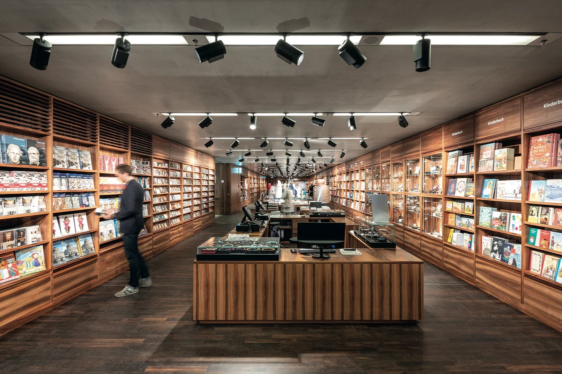 A bookstore with a warm wooden interior, illuminated by a combination of ceiling spotlights and linear light strips. Shelves full of books line the walls, while a central sales table organizes the room.