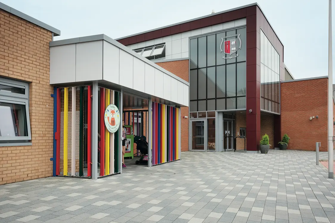 L'area esterna di una scuola con un tetto colorato fatto di montanti verticali. L'edificio moderno coniuga accenti rossi e facciate in vetro, mentre il cortile pavimentato offre un'atmosfera accogliente.