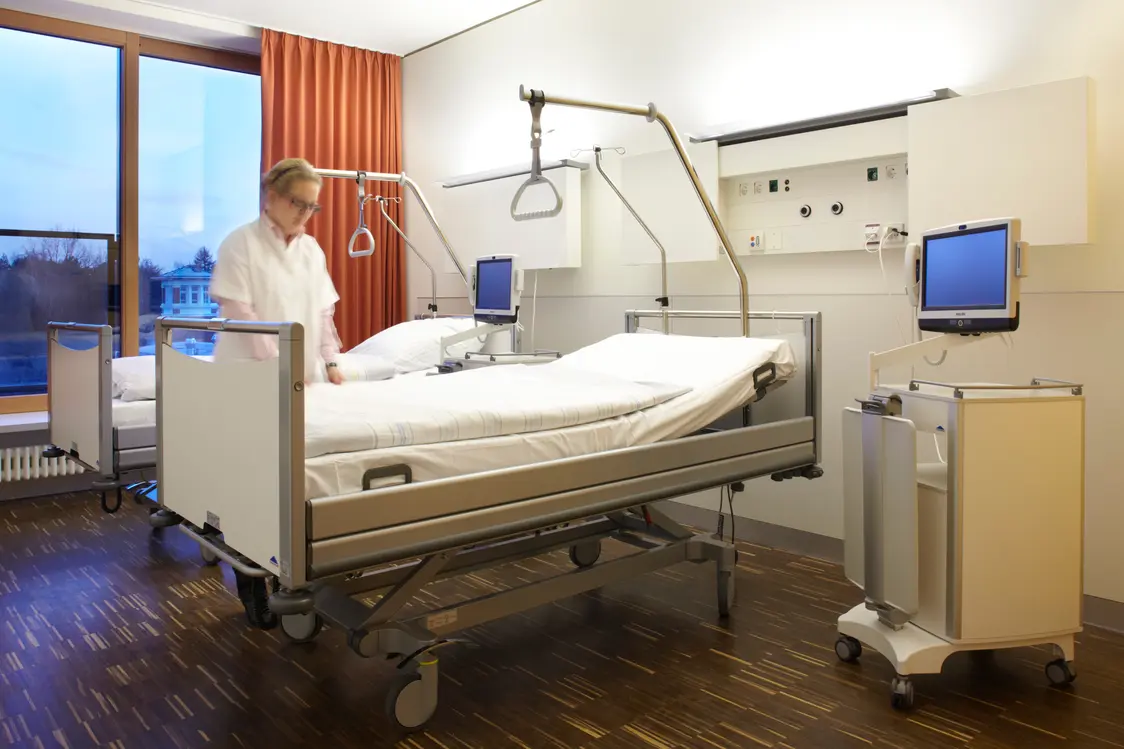 Ein Patientenzimmer mit zwei modernen Krankenhausbetten, medizinischer Ausstattung und einem großen Fenster, das Tageslicht hereinlässt. Die Beleuchtung ist dezent und sorgt für eine freundliche Atmosphäre.