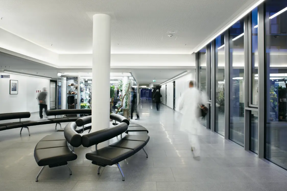 Ein heller Krankenhausflur mit großen Fensterfronten, modernen Sitzbänken und Pflanzen, gleichmäßig beleuchtet durch indirekte LED-Lichtbänder an Decke und Wänden. Personen in weißer Kleidung gehen durch den Flur.