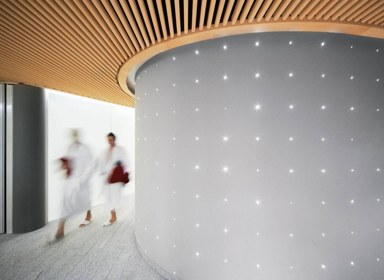  An interior of a wellness area with two people in bathrobes going through a brightly lit corridor. The wall is adorned with small lights, and the ceiling is covered with wooden panels.