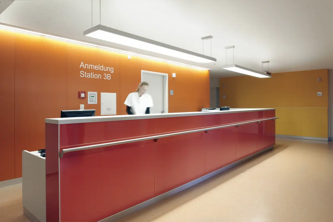 Ein Empfangsbereich mit langer, roter Theke und orangener Wand, beleuchtet durch zwei rechteckige Pendelleuchten. Eine Person in weißer Kleidung steht hinter dem Tresen, der Raum wirkt hell und freundlich.