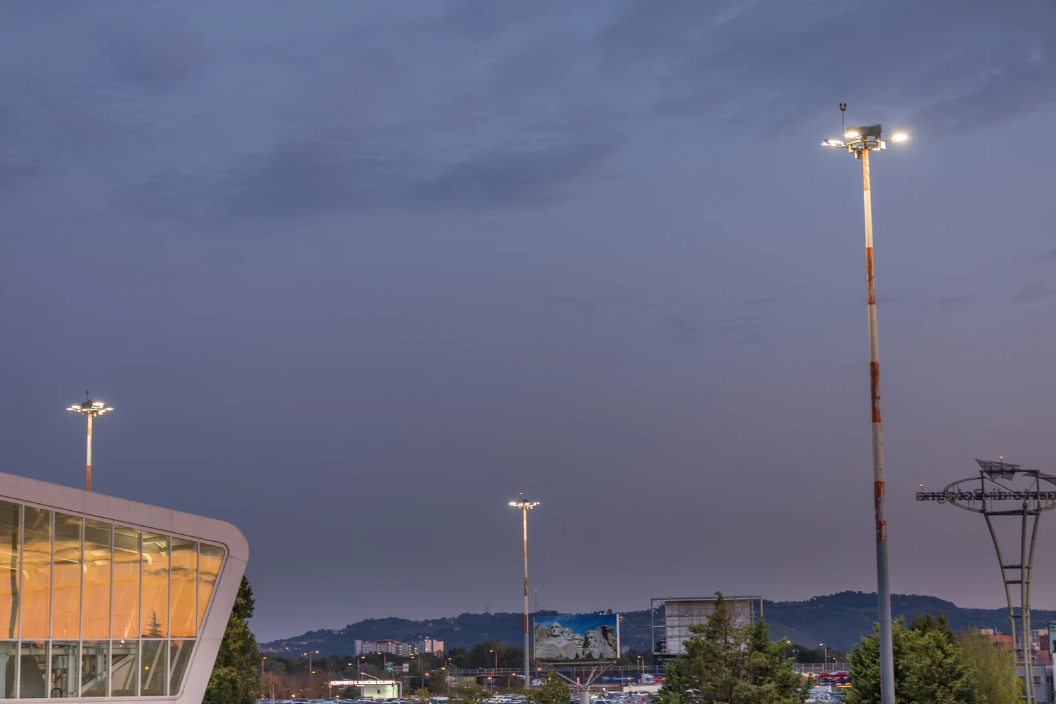 Buitenruimte bij schemering met hoge schijnwerpers die het tafereel verlichten. Op de achtergrond zijn stadsgebouwen en heuvels zichtbaar onder een heldere avondhemel.