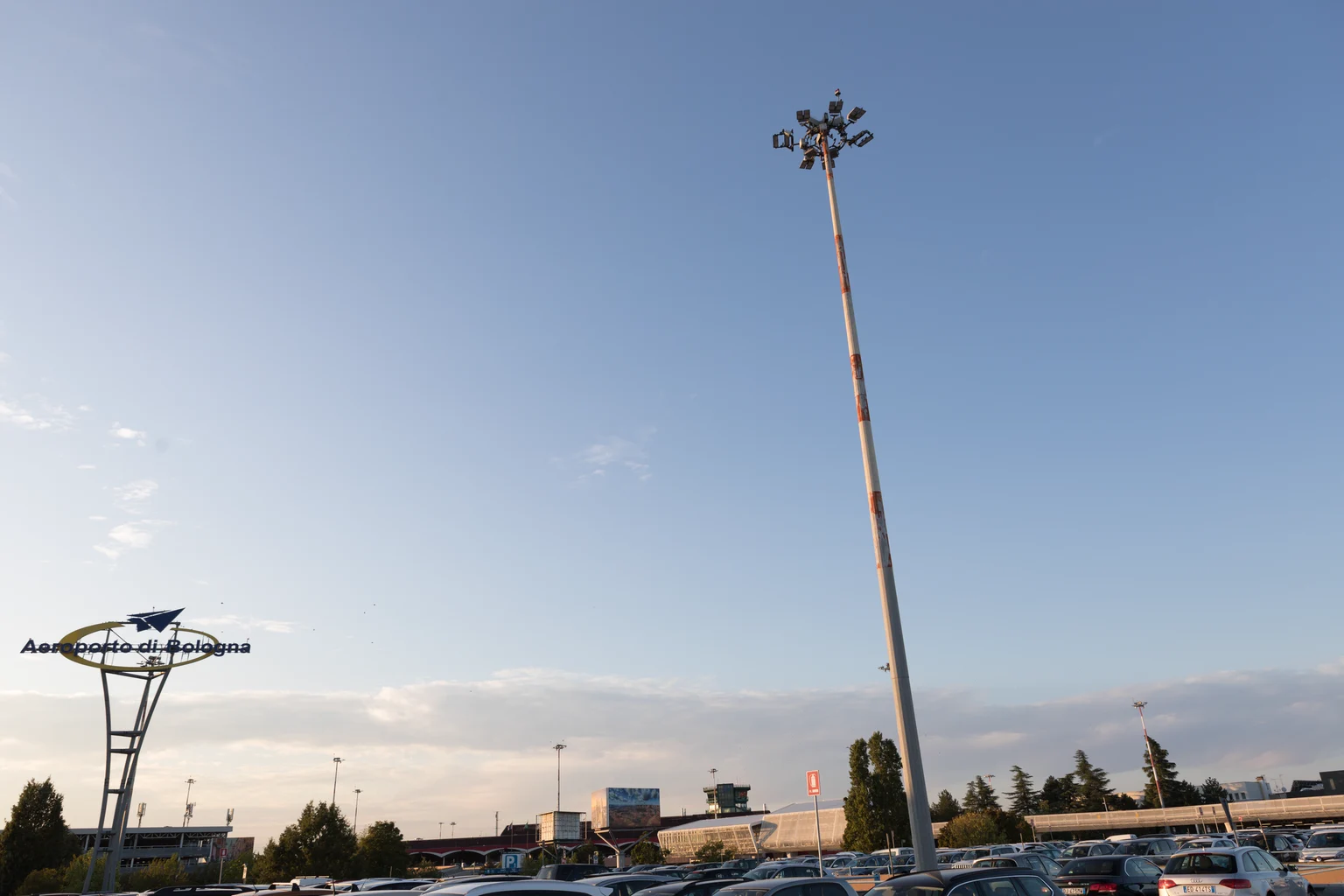 Grote parkeerplaats buiten met een hoge lichtmast in het midden. Onder een strakblauwe hemel zijn een aantal geparkeerde auto's en een gebouw zichtbaar.