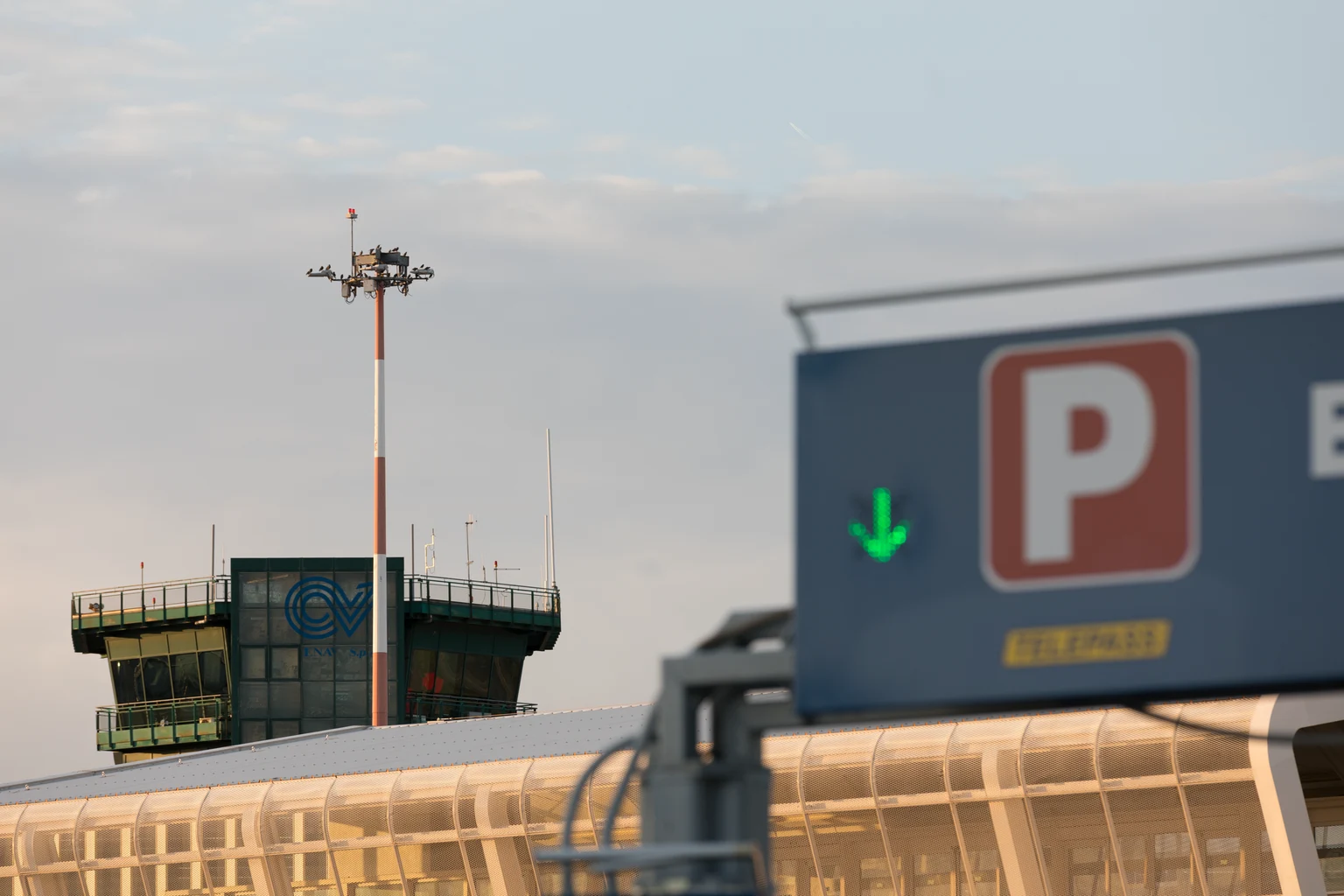 Verkeerstoren en terminalgebouw op de luchthaven bij zonsondergang, met een parkeerbord en een groene pijl op de voorgrond. De scène wordt zacht verlicht met een heldere hemel erboven.