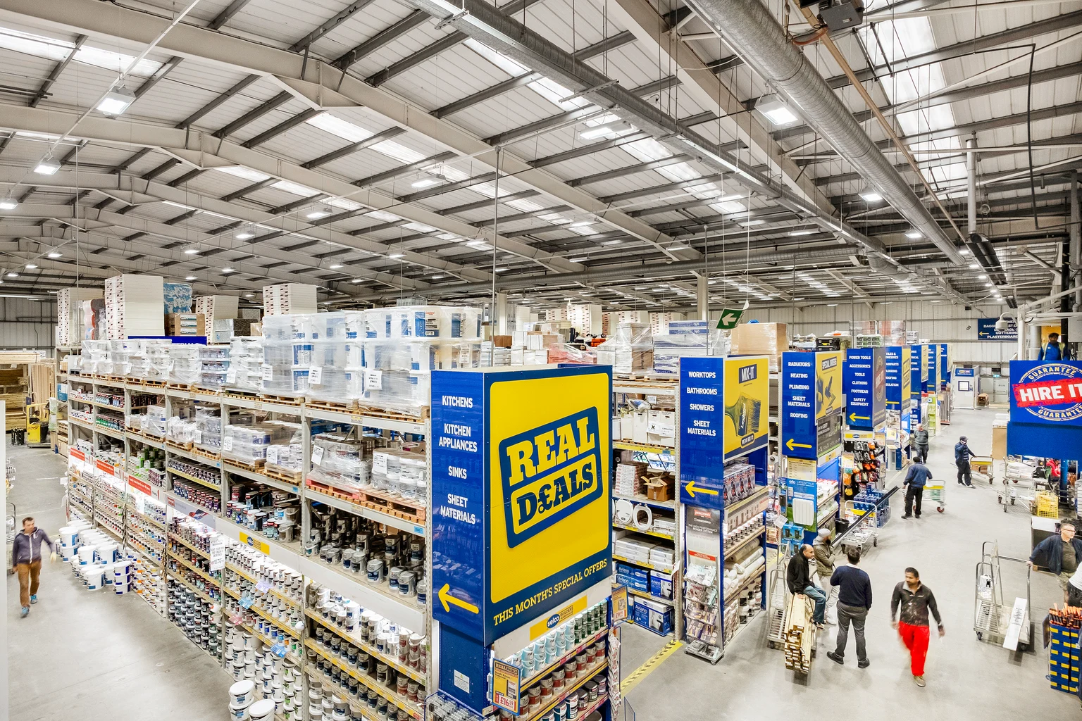 Een ruime bouwmarkt met hoge schappen waar verschillende producten zijn uitgestald, zoals gereedschap, verf en bouwmaterialen. Heldere verlichting en duidelijke borden leiden klanten door de georganiseerde gangpaden.