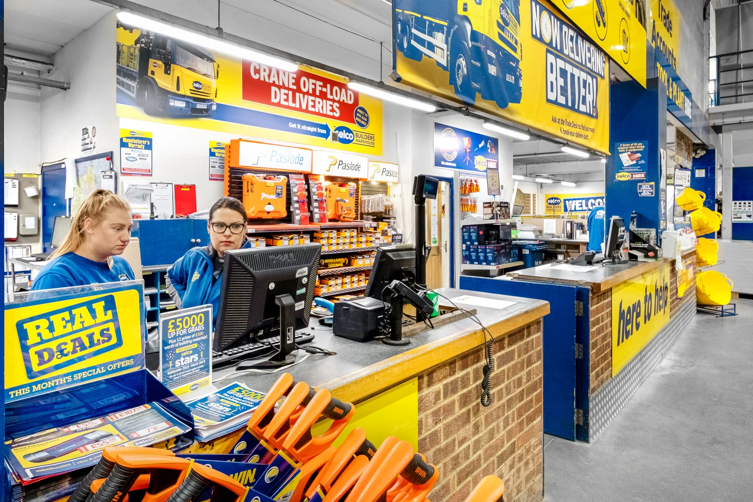 Een goed verlichte toonbank met promotiemateriaal en gereedschap op de voorgrond, omringd door felgele en blauwe branding. Twee medewerkers helpen klanten in een professionele en georganiseerde omgeving.