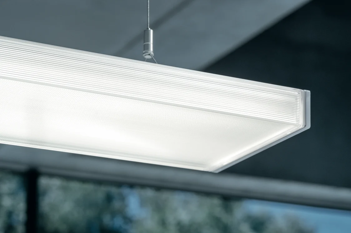 The ECOOS LED pendant luminaire emits evenly bright light in a modern room with large windows, providing optimal illumination.