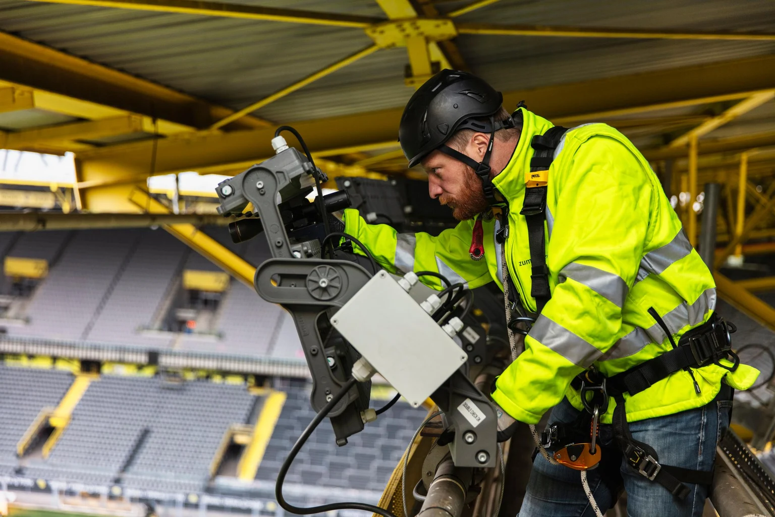 Een technicus in een opvallend veiligheidsvest en veiligheidsuitrusting werkt aan apparatuur in Signal Iduna Park. De iconische gele staalconstructies en de tribunes van het stadion zijn zichtbaar op de achtergrond.