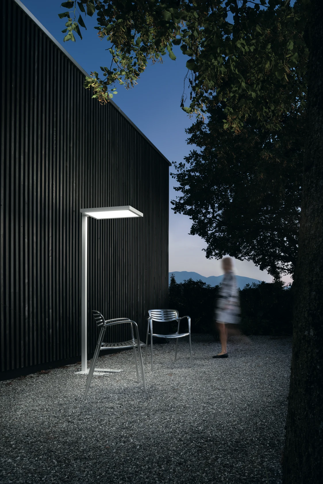 Le système d’éclairage EPURIA illumine une surface extérieure moderne avec deux chaises devant une façade en bois sombre au crépuscule.