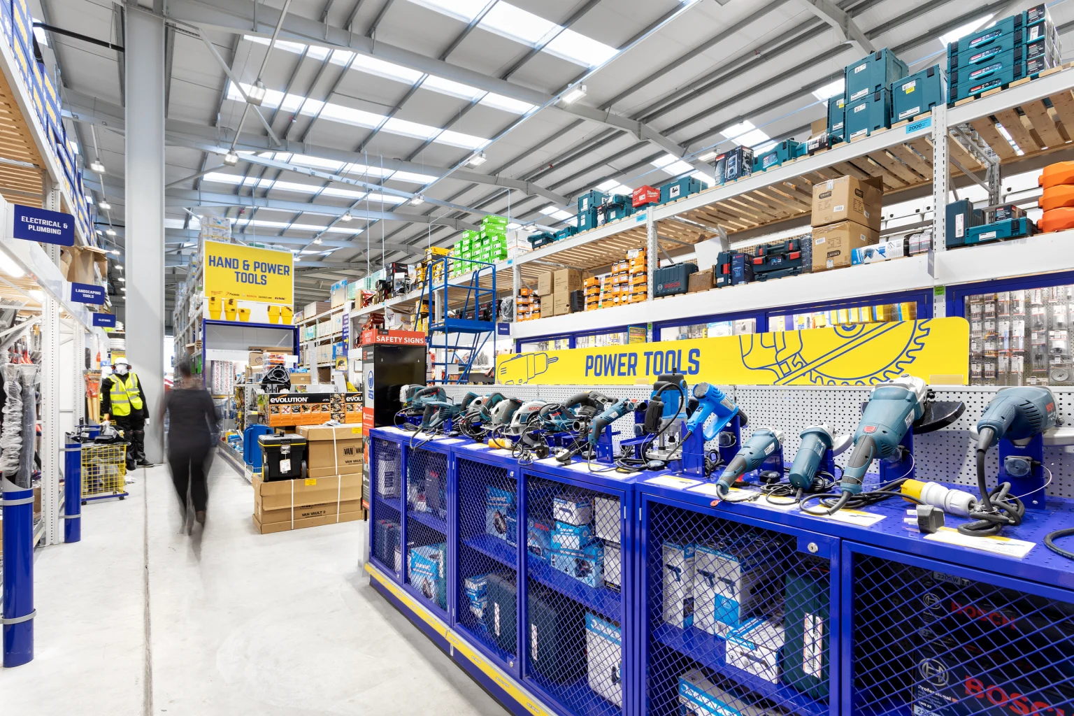 Een helder verlicht gangpad in een bouwmarkt met een verscheidenheid aan elektrisch gereedschap op blauwe schappen, met extra producten opgestapeld op hoge stellingen. Iemand loopt door de ruime, georganiseerde ruimte onder een plafond met dakraam.