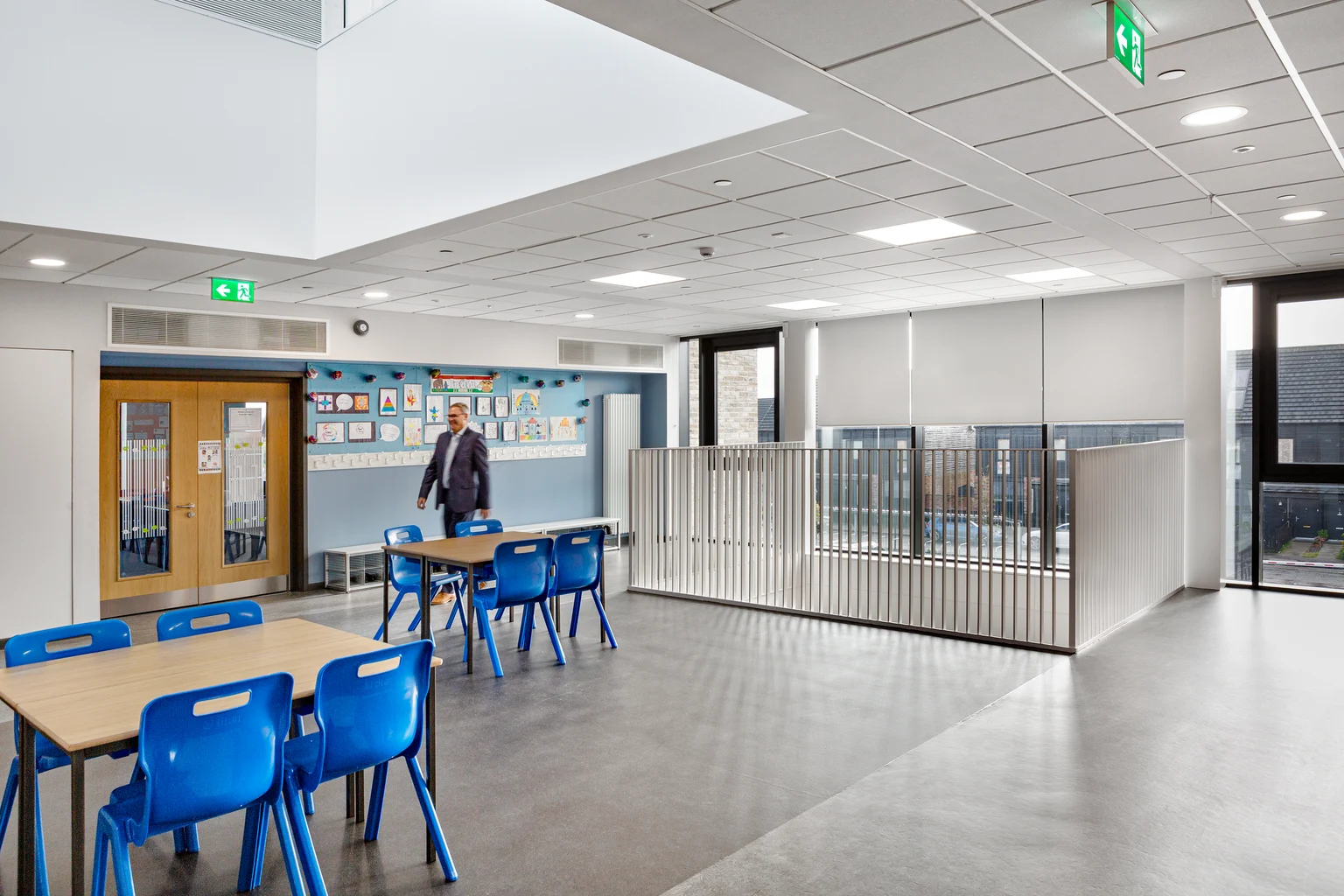 Un environnement scolaire lumineux et spacieux avec des chaises bleues et des tables en bois, baigné de lumière naturelle grâce à de grandes fenêtres. La pièce est décorée d’affiches éducatives aux murs et dispose d’un éclairage de plafond moderne pour un aspect propre et organisé.